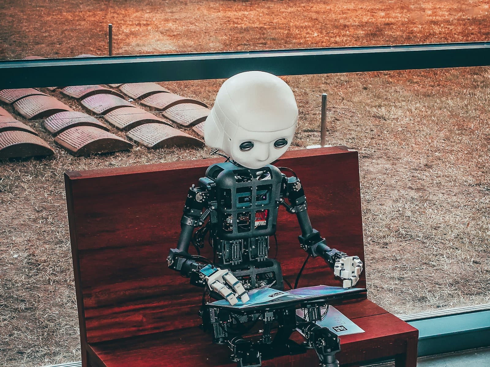 A small humanoid robot sitting on a wooden bench, reading a booklet