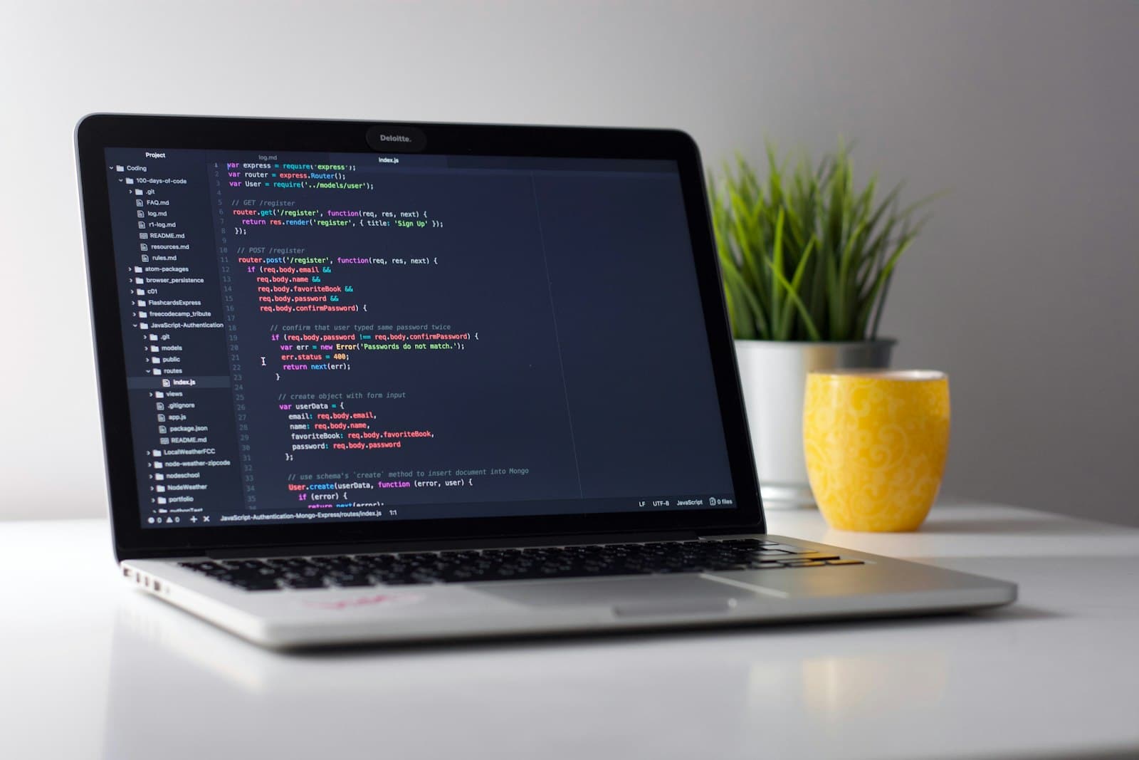 A laptop showing code on a bright desk beside a plant and a yellow mug