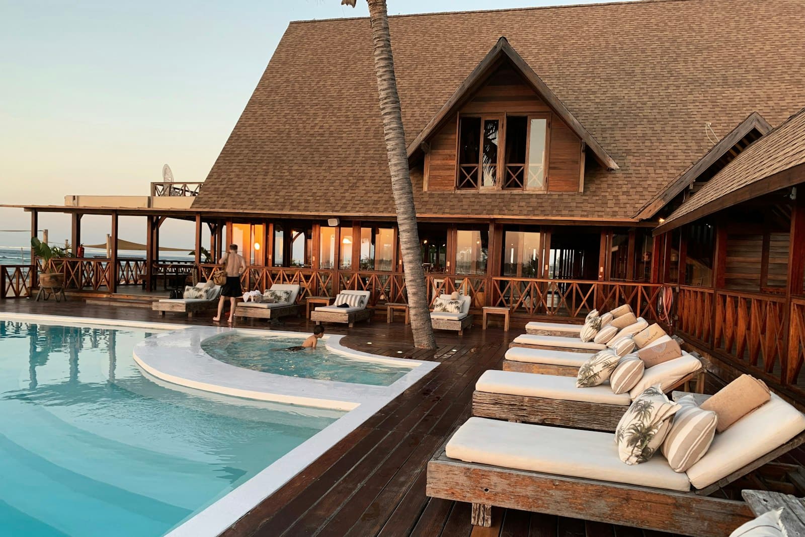 A resort pool with sun loungers in front of a wooden lodge at sunset